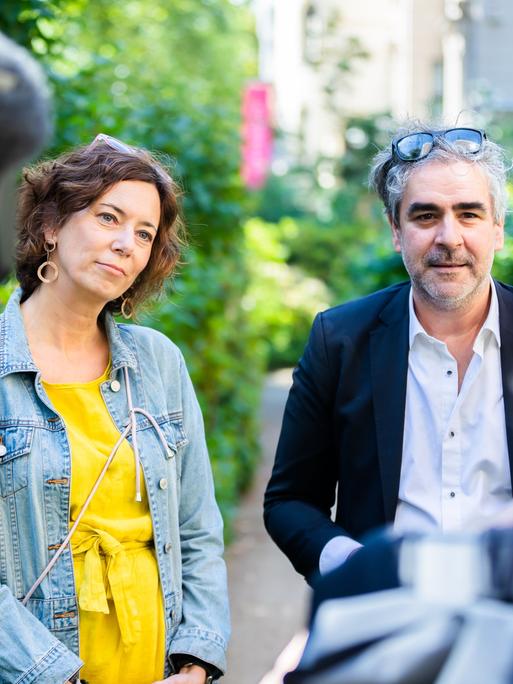 Eva Menasse und Deniz Yücel sprechen im Garten des Literaturhaus Berlin mit Journalisten. Sie trägt ein gelbes Kleid und eine Jeansjacke, er ein helles Hemd und Sakko. Beide haben Brillen ins Haar hochgeschoben. Links ist unscharf ein Mann mit Kamera auf der Schulter von hinten zu sehen, rechts der Hinterkopf und die Schulter einer Frau.