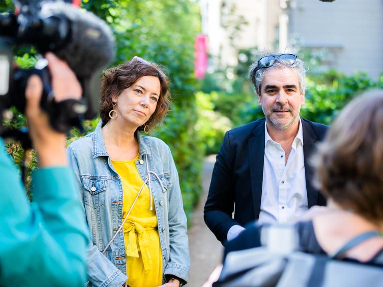 Eva Menasse und Deniz Yücel sprechen im Garten des Literaturhaus Berlin mit Journalisten. Sie trägt ein gelbes Kleid und eine Jeansjacke, er ein helles Hemd und Sakko. Beide haben Brillen ins Haar hochgeschoben. Links ist unscharf ein Mann mit Kamera auf der Schulter von hinten zu sehen, rechts der Hinterkopf und die Schulter einer Frau.