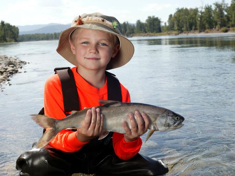 Ein Junge mit Anglerhütchen hält stolz einen Fisch in die Kamera, im Hintergrund sieht man einen See