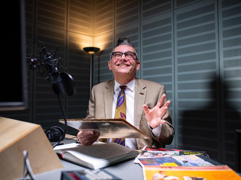 Moderator Götz Alsmann sitzt im WDR-Tonstudio in Münster vor dem Mikrofon und hält ein altes Plattencover in der Hand.