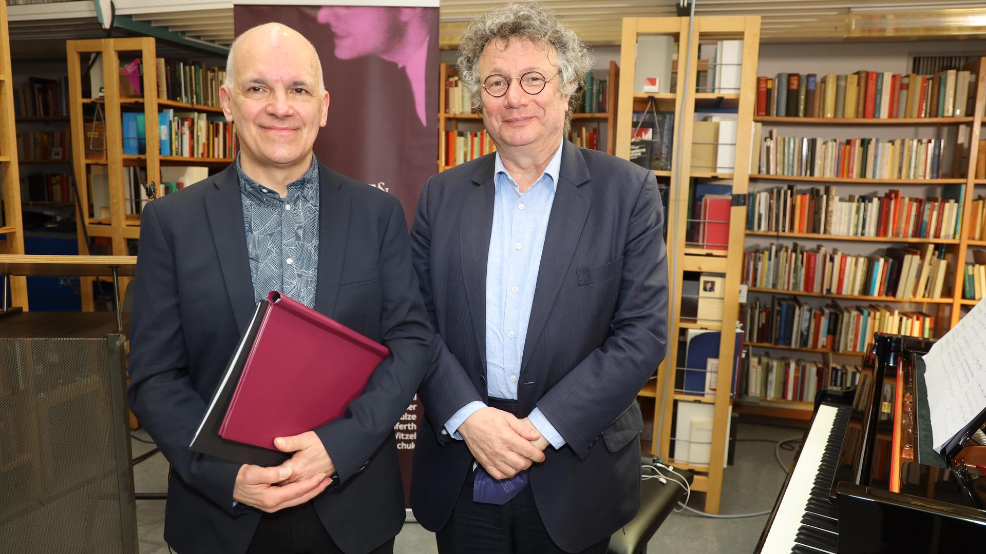 Die Schriftsteller Frank Witzel und Ingo Schulze bei der Verleihung des Erich-Fried-Preises 2021. Witzel hält eine Mappe in der Hand, hinter ihnen sind Regalreihen voller Bücher, neben ihnen ein Klavier.