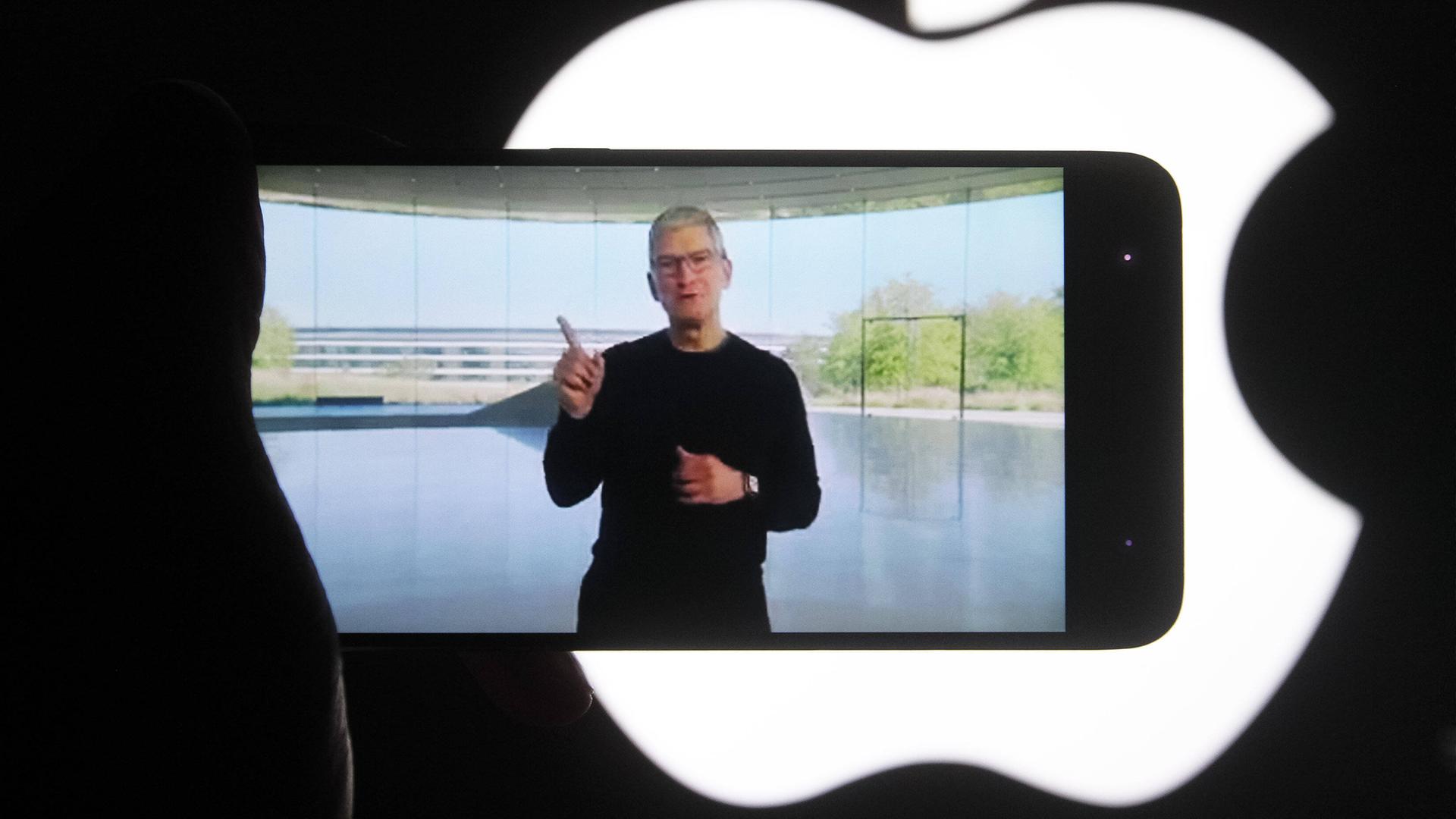 Der bisherige Apple Chef Tim Cook ist auf dem Bildschirm eines iPhones zu sehen. Im Hintergrund sieht man das Apple-Logo (Archivbild).