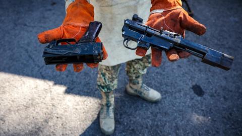 Eine Person in Militäruniform hält zwei Waffen mit abgesägten Läufen in die Kamera.
