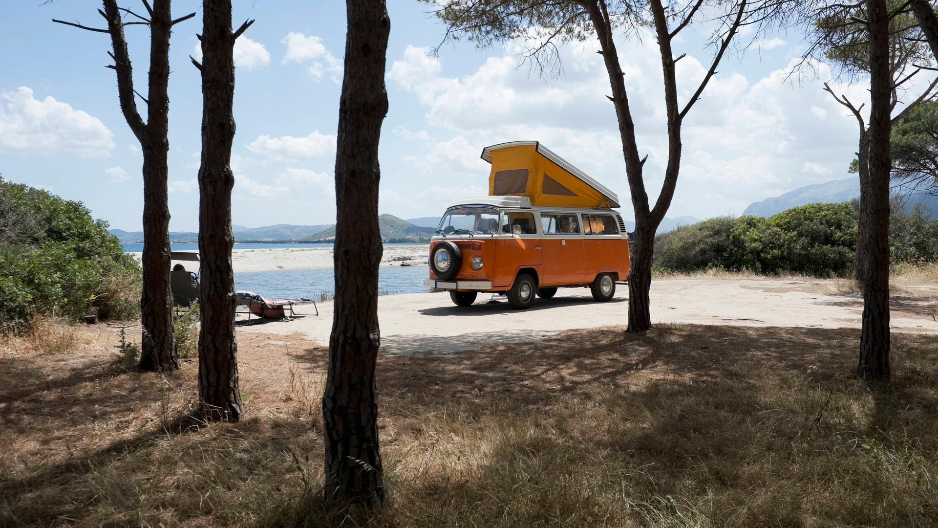 Ein alter orangener VW Bus steht mit ausgeklapptem Dachzelt zwischen Bäumen an einer Küste auf Sardinien.