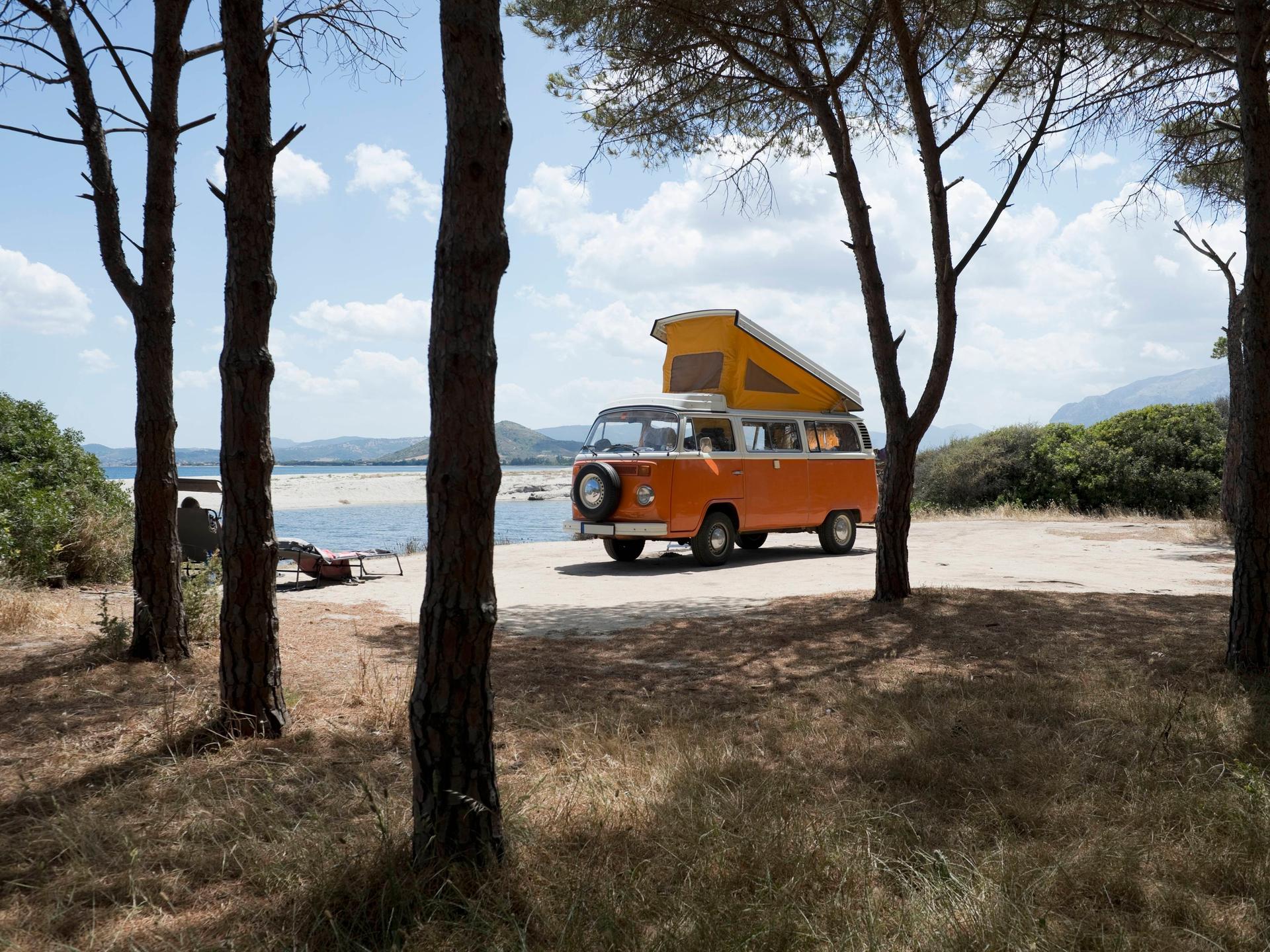 Ein alter orangener VW Bus steht mit ausgeklapptem Dachzelt zwischen Bäumen an einer Küste auf Sardinien.