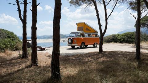Ein alter orangener VW Bus steht mit ausgeklapptem Dachzelt zwischen Bäumen an einer Küste auf Sardinien.
