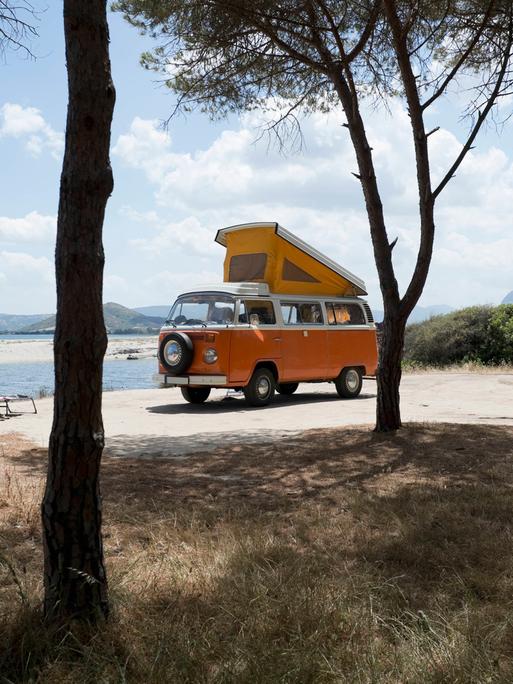 Ein alter orangener VW Bus steht mit ausgeklapptem Dachzelt zwischen Bäumen an einer Küste auf Sardinien.