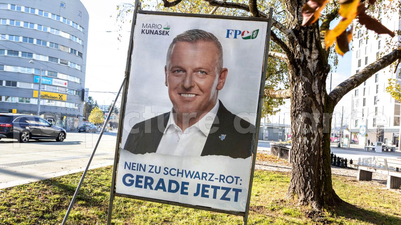 Das Plakat zeigt den lächelnden Politiker. Es steht auf einem Grünstreifen zwischen zwei Fahrbahnen einer Hauptverkehrsstraße. Dahinter Gebäude.