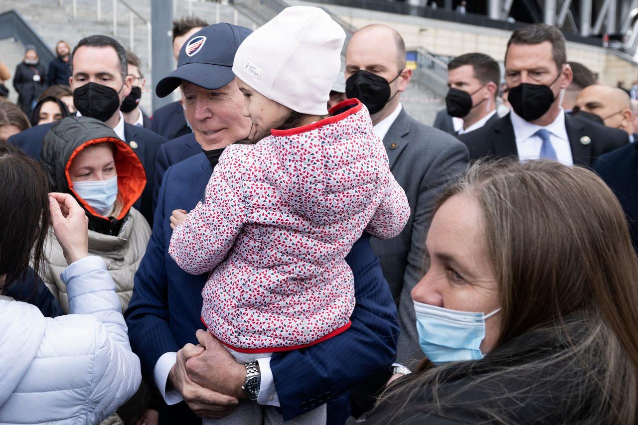 US-Präsident Biden mit einem Mädchen auf dem Arm, umringt von Flüchtlingen und Bodyguards mit Mundschutz.