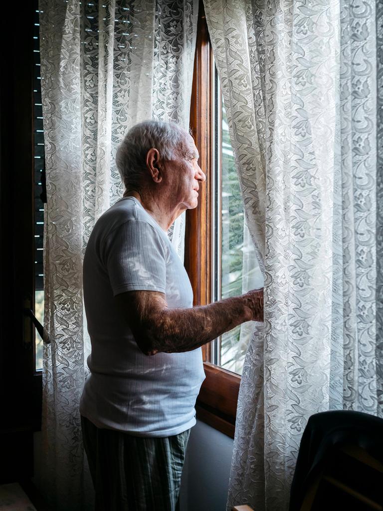 Ein alter Mann schaut aus seinem Fenster und schiebt dabei die Spitzengardine zur Seite.