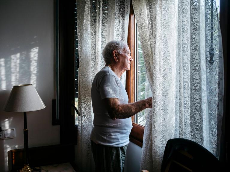 Ein alter Mann schaut aus seinem Fenster und schiebt dabei die Spitzengardine zur Seite.