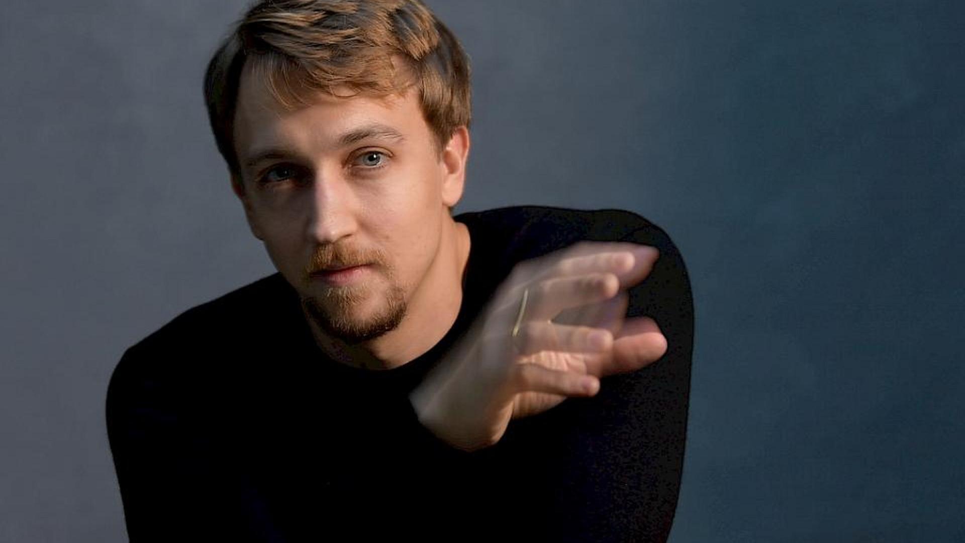 Fabian Müller sitzt im schlicht-schwarzen Pullover vor der Kamera und scheint mit einer Hand zu dirigieren, die etwas verwischt erscheint. Fabian Müller sitzt im schlicht-schwarzen Pullover vor der Kamera und scheint mit einer Hand zu dirigieren, die etwas verwischt erscheint.