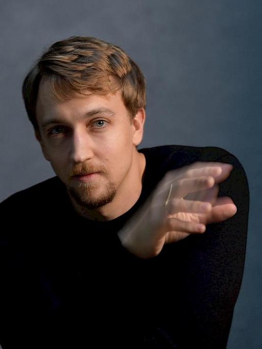 Fabian Müller sitzt im schlicht-schwarzen Pullover vor der Kamera und scheint mit einer Hand zu dirigieren, die etwas verwischt erscheint.