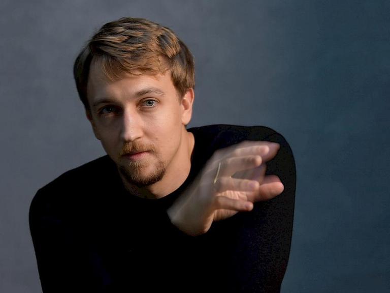 Fabian Müller sitzt im schlicht-schwarzen Pullover vor der Kamera und scheint mit einer Hand zu dirigieren, die etwas verwischt erscheint.