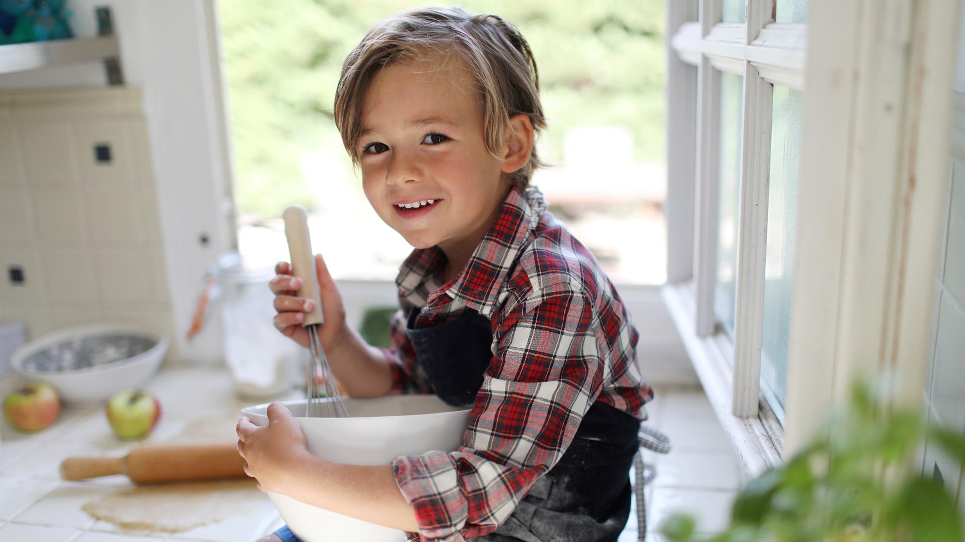 Ein ca. 4-5 jähriger bloinder junge schaut lachend in die Kamera, während er in einer Rührschüssel rührt