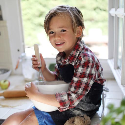 Ein ca. 4-5 jähriger bloinder junge schaut lachend in die Kamera, während er in einer Rührschüssel rührt
