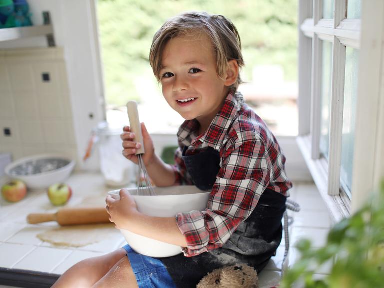 Ein ca. 4-5 jähriger bloinder junge schaut lachend in die Kamera, während er in einer Rührschüssel rührt