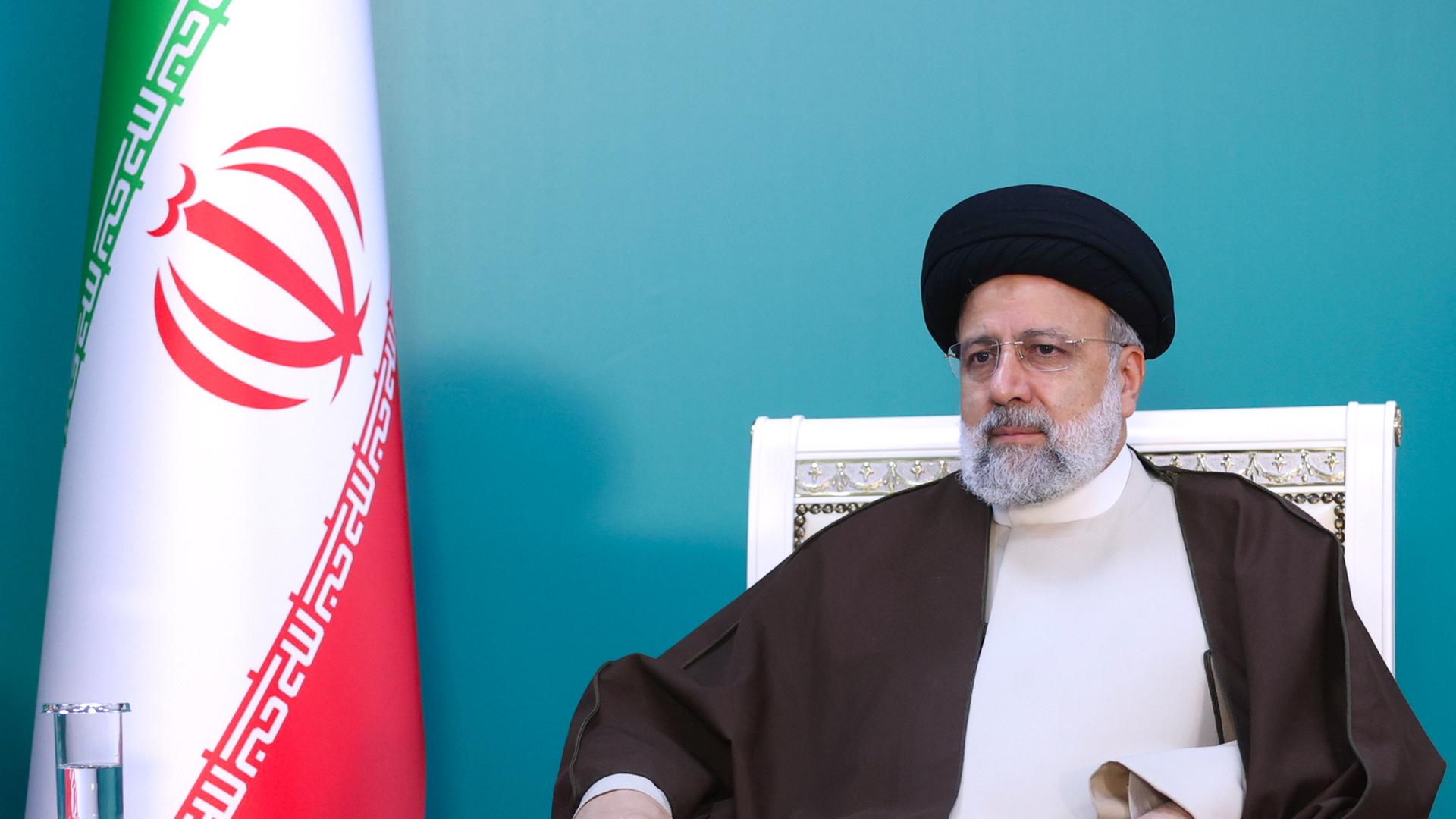 Präsident Ebrahim Raisi sitzt auf einem weißen Stuhl. Recht neben ihm ist die iranische Flagge zu sehen.