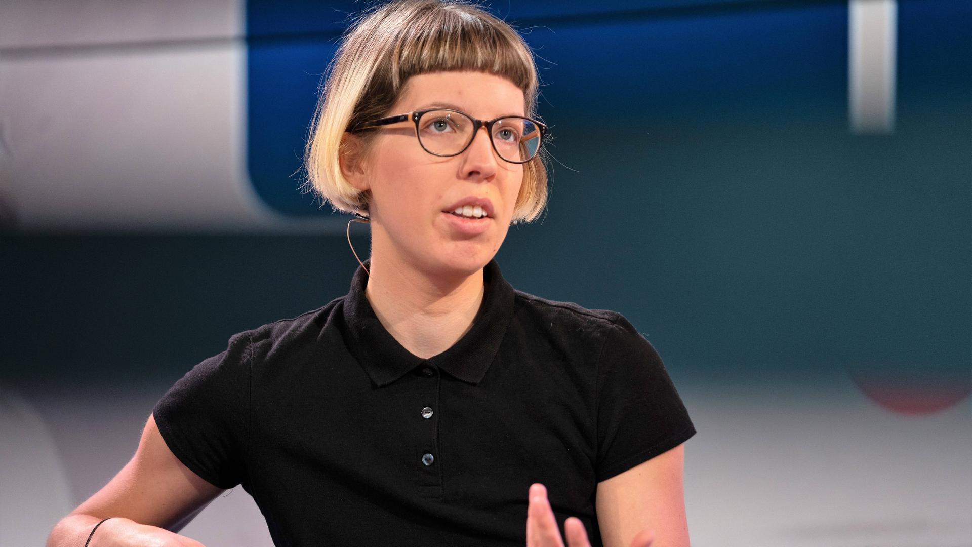 Die Autorin Bettina Wilpert sitzt auf einem blauen Sessel und gestikuliert, sie trägt reine schwarze Brille und ein schwarzes T-Shirt.