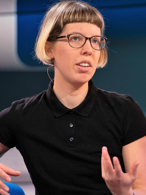 Die Autorin Bettina Wilpert sitzt auf einem blauen Sessel und gestikuliert, sie trägt reine schwarze Brille und ein schwarzes T-Shirt.