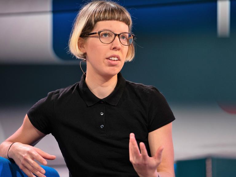 Die Autorin Bettina Wilpert sitzt auf einem blauen Sessel und gestikuliert, sie trägt reine schwarze Brille und ein schwarzes T-Shirt.