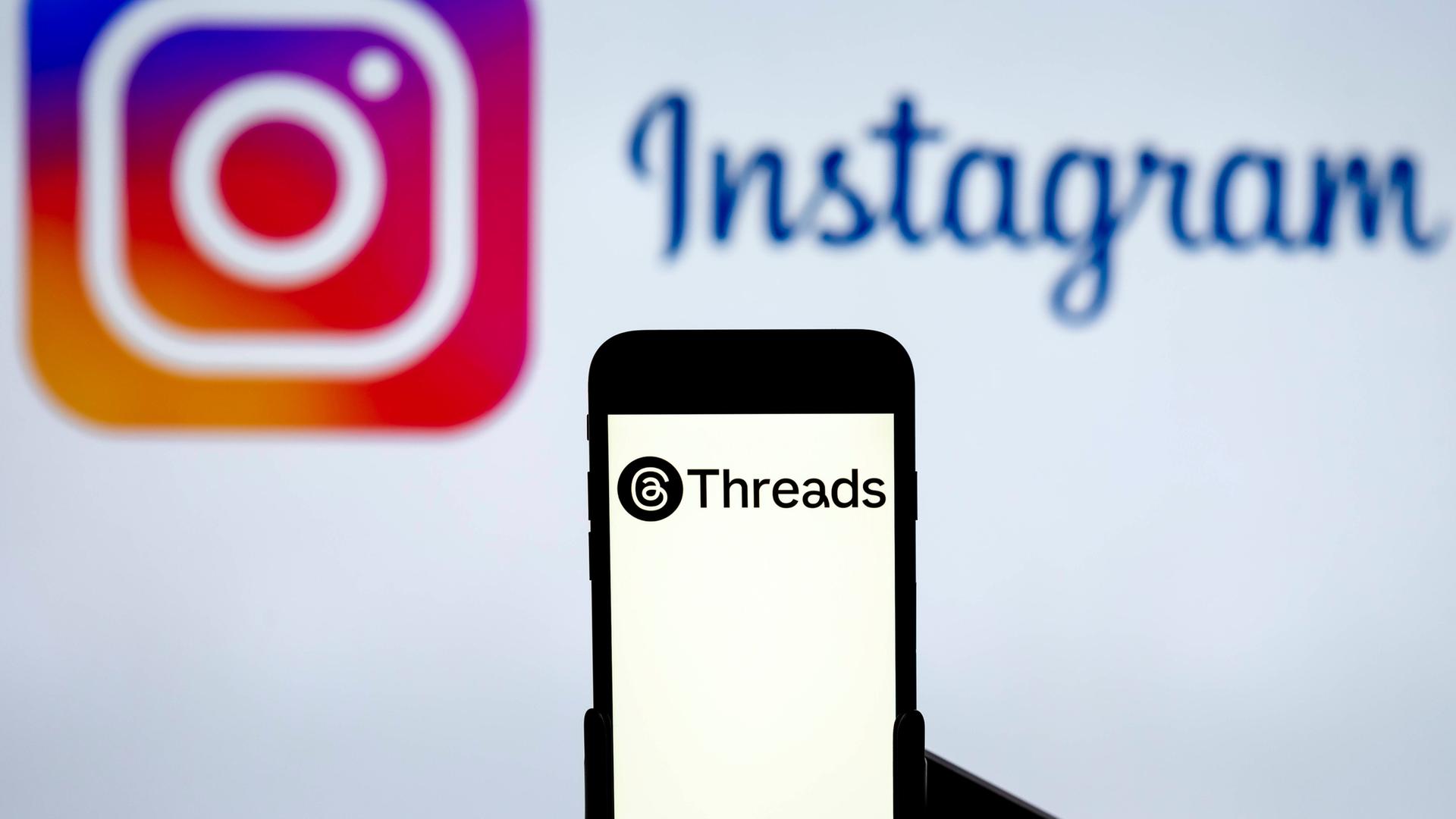 Ein Handy mit dem Logo des Dienstes Threads, im Hintergrund das Logo von Instagram auf einem Computerbildschirm