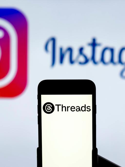 Ein Handy mit dem Logo des Dienstes Threads, im Hintergrund das Logo von Instagram auf einem Computerbildschirm