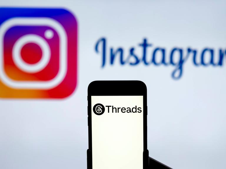 Ein Handy mit dem Logo des Dienstes Threads, im Hintergrund das Logo von Instagram auf einem Computerbildschirm