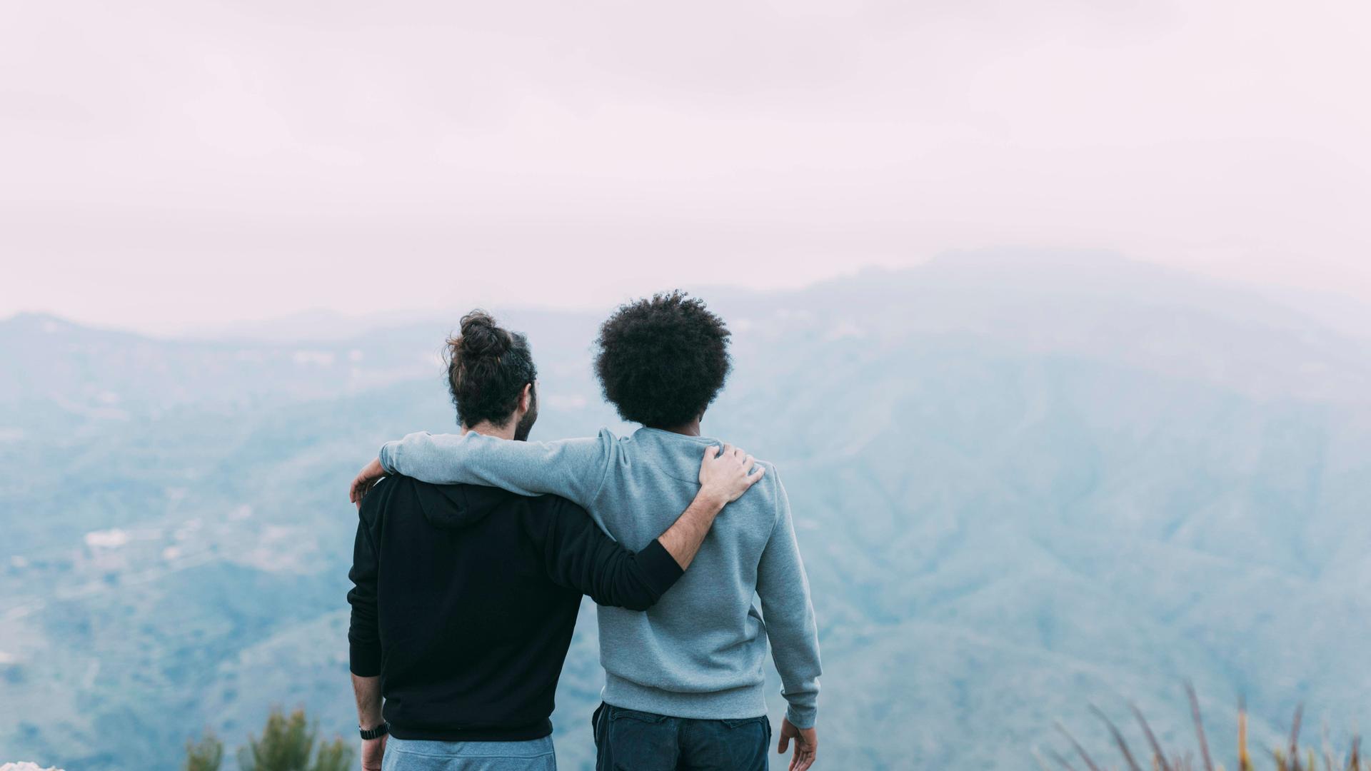 Zwei Freunde stehen Arm in Arm mit dem Rücken zur Kamera auf einem Berg und schauen in Richtung Horizont.