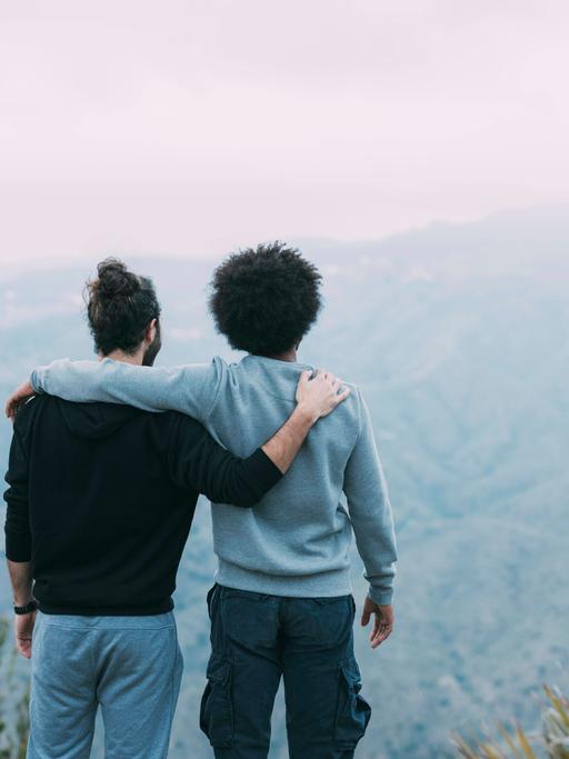 Zwei Freunde stehen Arm in Arm mit dem Rücken zur Kamera auf einem Berg und schauen in Richtung Horizont.