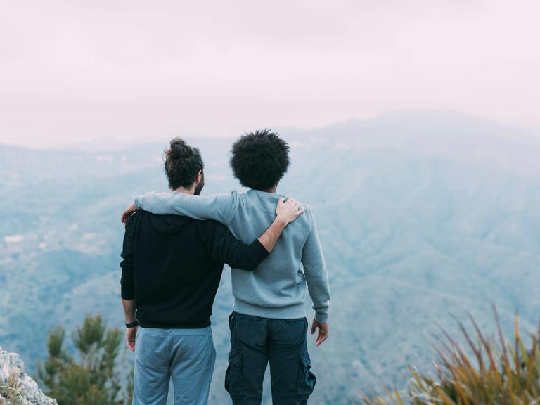 Zwei Freunde stehen Arm in Arm mit dem Rücken zur Kamera auf einem Berg und schauen in Richtung Horizont.