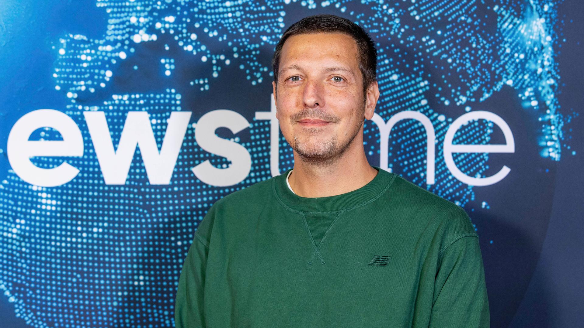 Thilo Mischke beim newstime Empfang der ProSiebenSat.1-Poltikredaktion im Hauptstadtbüro der Seven.One Entertainment Group. Er trägt einen dunkelgrünen Pullover und steht vor einem blauen Hintergrund.