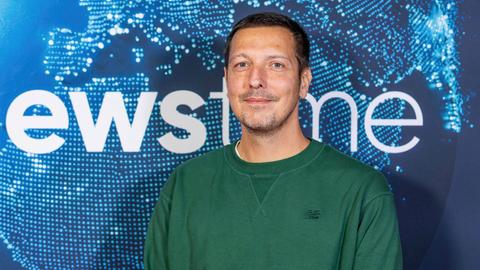 Thilo Mischke beim newstime Empfang der ProSiebenSat.1-Poltikredaktion im Hauptstadtbüro der Seven.One Entertainment Group. Er trägt einen dunkelgrünen Pullover und steht vor einem blauen Hintergrund.