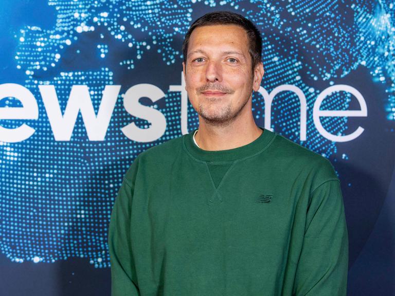 Thilo Mischke beim newstime Empfang der ProSiebenSat.1-Poltikredaktion im Hauptstadtbüro der Seven.One Entertainment Group. Er trägt einen dunkelgrünen Pullover und steht vor einem blauen Hintergrund.