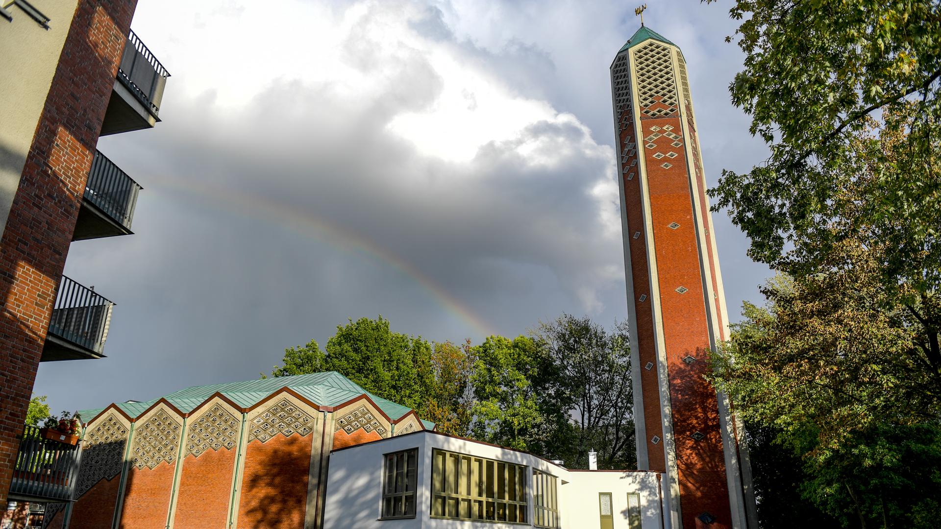 Außenansicht einer ehemaligen evangelischen Kirche aus den 1960er-Jahren mit einem schlanken Backstein-Turm, auf dem jedoch kein Kreuz mehr angebracht ist. Im Hintergrund: ein Regenbogen.