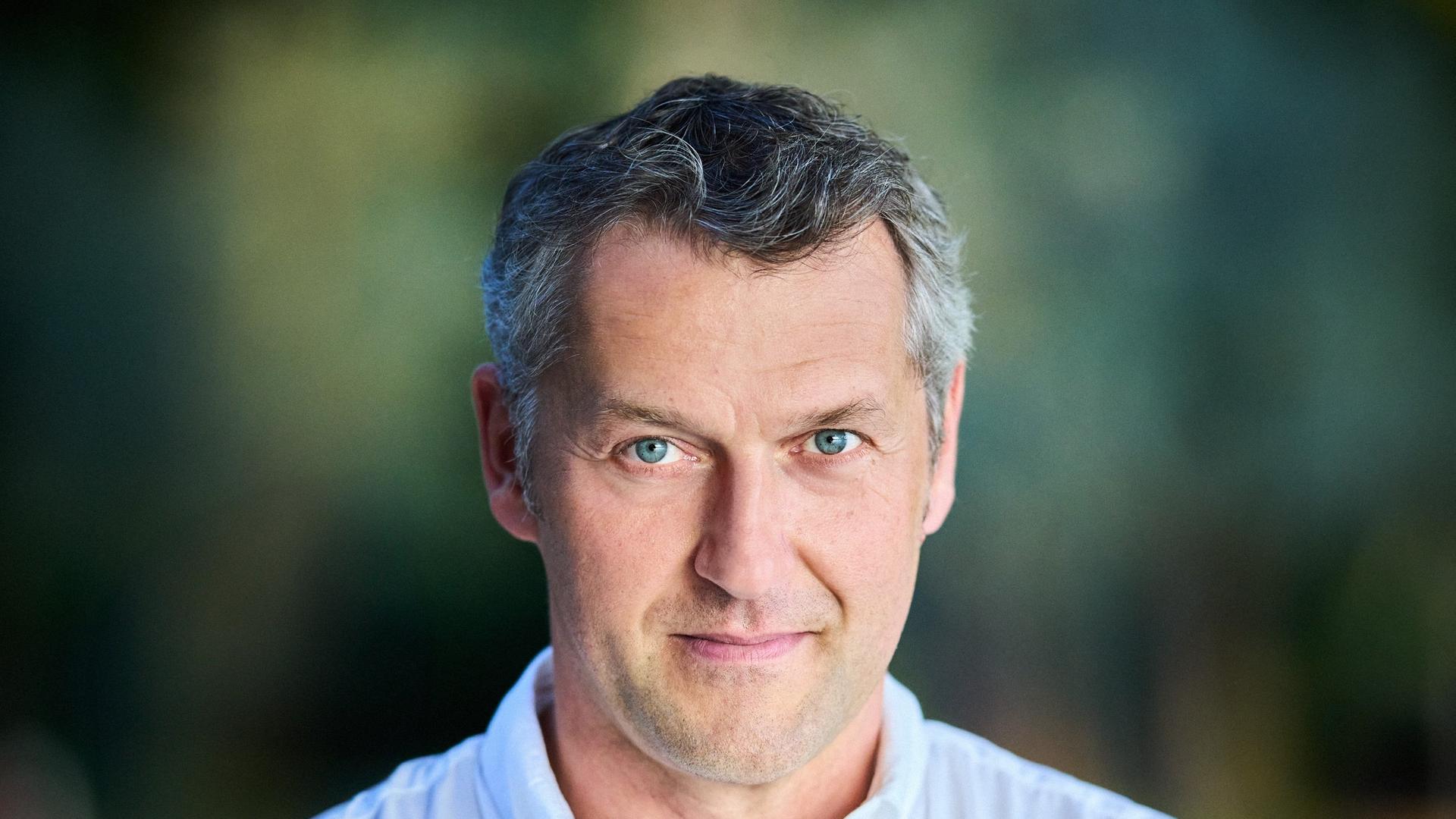 Ein Mann mit grauem Haar und blauen Augen schaut freundlich in die Kamera. Er trägt ein hellblaues Hemd, hinter ihm ist verschwommen Grün zu sehen. Er verschränkt die Arme vor dem Oberkörper. Ein Mann mit grauem Haar und blauen Augen schaut freundlich in die Kamera. Er trägt ein hellblaues Hemd, hinter ihm ist verschwommen Grün zu sehen. Er verschränkt die Arme vor dem Oberkörper.