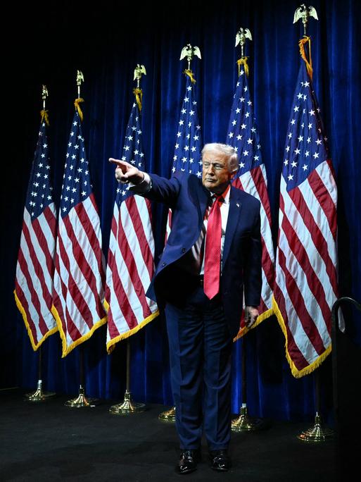 Donald Trump steht auf einer Bühne vor US-Flaggen und zeigt mit ausgestrecktem Arm und Zeigefinger ins Publikum.
