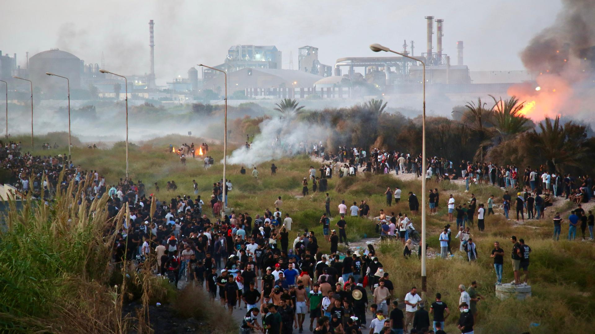 Blick von einer Anhöhe auf Demonstranten auf einem Feld; die Szenerie ist verraucht. Hinter einem Gebüsch brennt ein Feuer. Im Hintergrund die Chemieanlage. Blick von einer Anhöhe auf Demonstranten auf einem Feld; die Szenerie ist verraucht. Hinter einem Gebüsch brennt ein Feuer. Im Hintergrund die Chemieanlage.