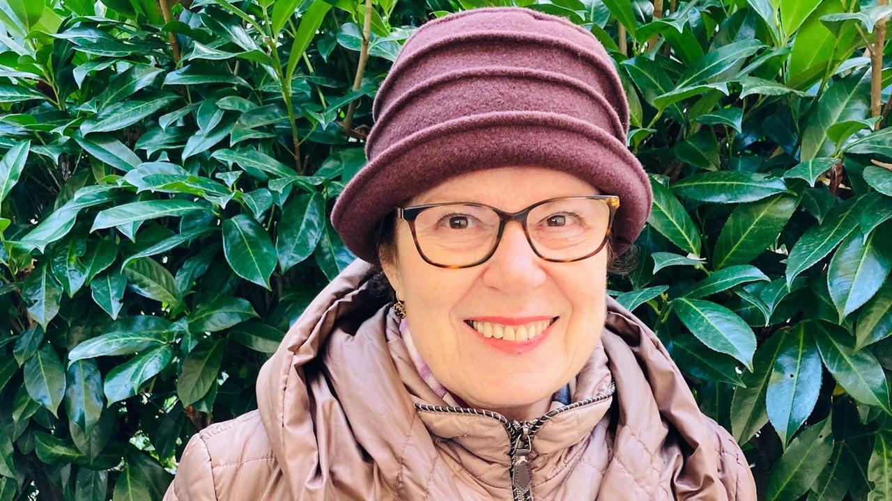 Porträt von Judith Landshut vor einem Busch mit grünen Blättern. Sie trägt eine Brille und einen Hut. Porträt von Judith Landshut vor einem Busch mit grünen Blättern. Sie trägt eine Brille und einen Hut.