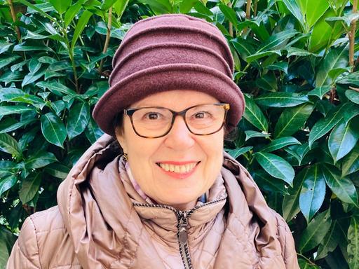 Porträt von Judith Landshut vor einem Busch mit grünen Blättern. Sie trägt eine Brille und einen Hut.