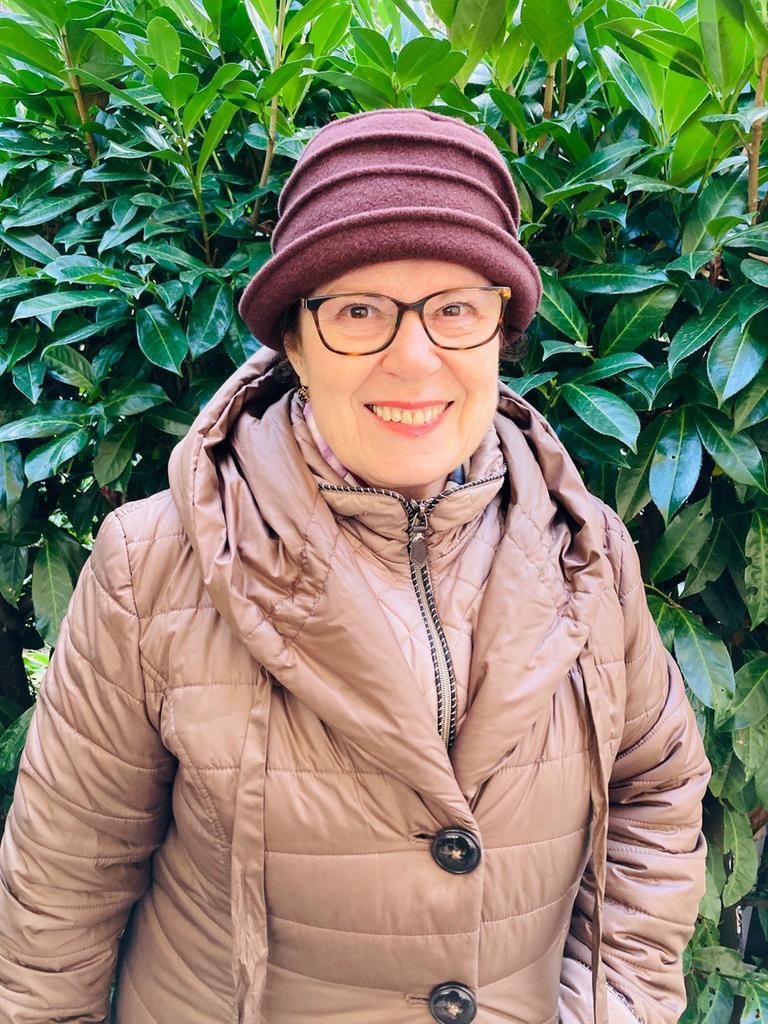 Porträt von Judith Landshut vor einem Busch mit grünen Blättern. Sie trägt eine Brille und einen Hut.