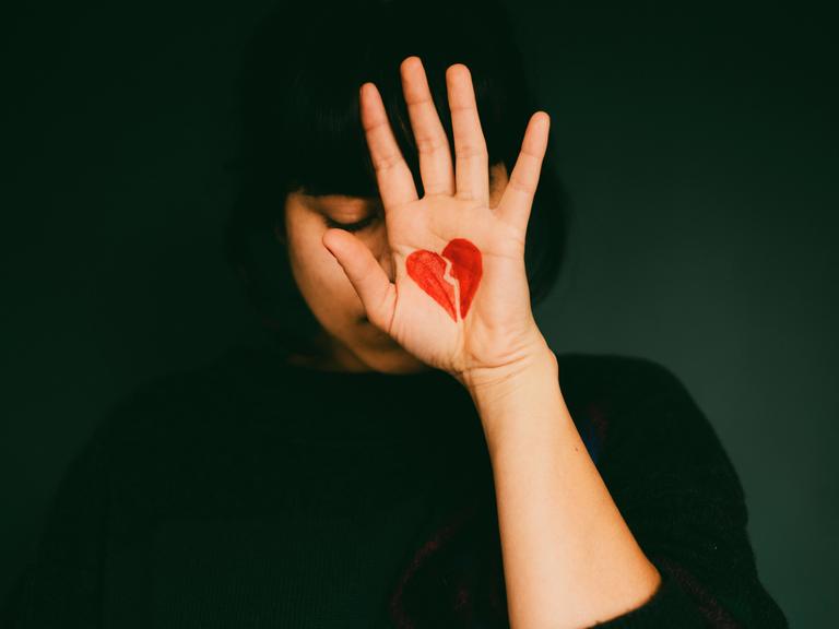 Frau hält sich die Hand, auf die ein zerbrochenes Herz gemalt ist, vor das Gesicht. Konzept Trennung und Liebeskummer.