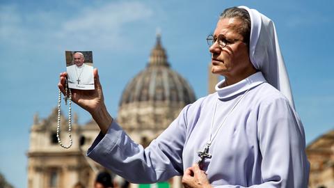 Eine Nonne in hellblauem Ornat hält ein Bild von Papst Franziskus und einen Rosenkranz in der Hand; im Hintergrund die Kuppel des Petersdoms.