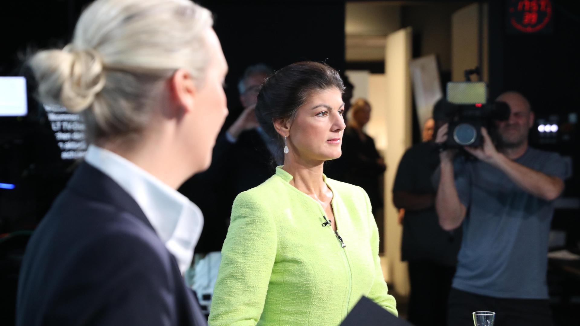 Alice Weidel steht seitlich mit ihrem Kopf von der Kamera weggedreht zu Sahra Wagenknecht rüber, die in einem gelbgrünen Sakko zufrieden nach vorn schaut. Das Foto wurde im Studio von Welt-TV aufgenommen.