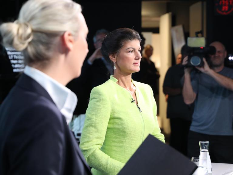 Alice Weidel steht seitlich mit ihrem Kopf von der Kamera weggedreht zu Sahra Wagenknecht rüber, die in einem gelbgrünen Sakko zufrieden nach vorn schaut. Das Foto wurde im Studio von Welt-TV aufgenommen.