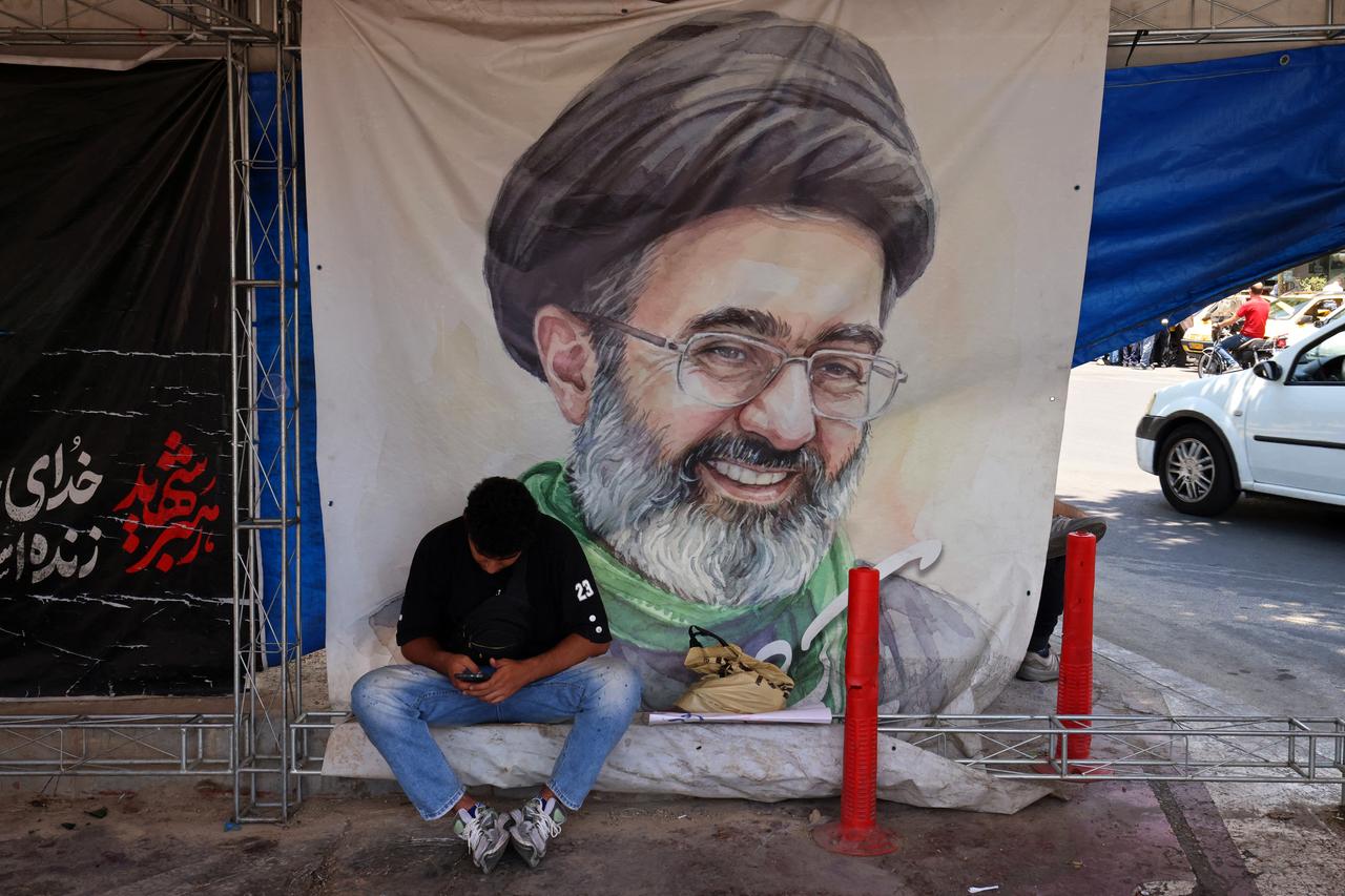 Ein Mann sitzt am 10. Juni 2026 auf dem Vanak-Platz in Teheran vor einem Plakat mit dem Porträt des iranischen Obersten Führers Mojtaba Khamenei, er schaut auf sein Mobiltelefon herab.