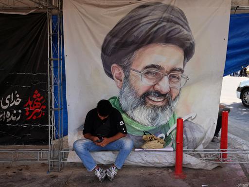 Ein Mann sitzt am 10. Juni 2026 auf dem Vanak-Platz in Teheran vor einem Plakat mit dem Porträt des iranischen Obersten Führers Mojtaba Khamenei, er schaut auf sein Mobiltelefon herab. Ein Mann sitzt am 10. Juni 2026 auf dem Vanak-Platz in Teheran vor einem Plakat mit dem Porträt des iranischen Obersten Führers Mojtaba Khamenei, er schaut auf sein Mobiltelefon herab.