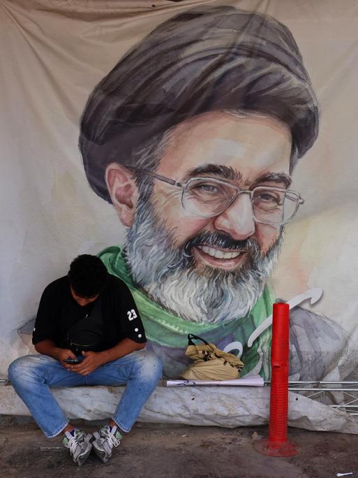 Ein Mann sitzt am 10. Juni 2026 auf dem Vanak-Platz in Teheran vor einem Plakat mit dem Porträt des iranischen Obersten Führers Mojtaba Khamenei, er schaut auf sein Mobiltelefon herab. Ein Mann sitzt am 10. Juni 2026 auf dem Vanak-Platz in Teheran vor einem Plakat mit dem Porträt des iranischen Obersten Führers Mojtaba Khamenei, er schaut auf sein Mobiltelefon herab.