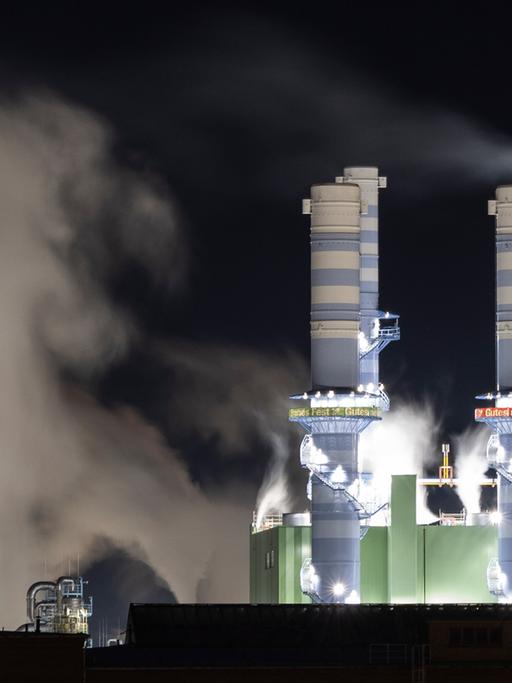 Dampf steigt bei Nacht aus beleuchteten Industrieschornsteinen des Industriepark Höchst auf. 