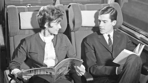 Zugreisen in den 1960er Jahren - ein Paar sitzt im Zug, die Frau eine Zeitschrift, der Mann ein Buch lesend.